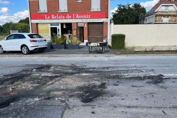 Restes de voiture brûlée sur la route, avec en arrière-plan le restaurant dont la vitrine a été cassée.