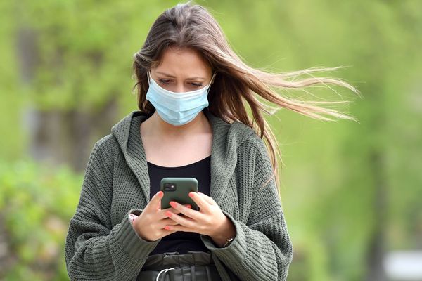 Une jeune femme portant un masque consulte son smartphone - Photo d'illustration