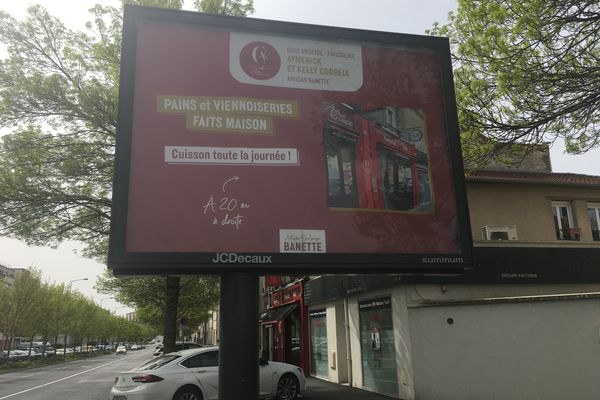 La boulangerie, qu'on devine avec sa devanture rouge, juste derrière le panneau publicitaire.